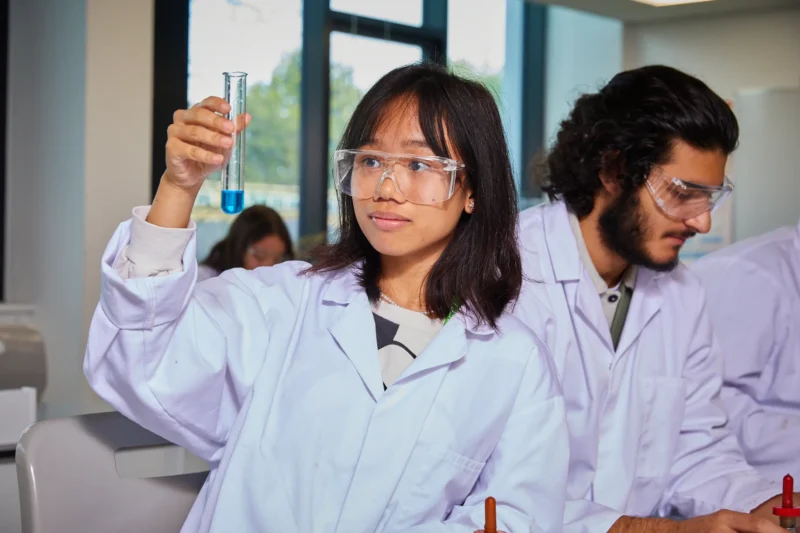 Science student wearing laboratory coat and safety glasses hold up test tube in science classroom.