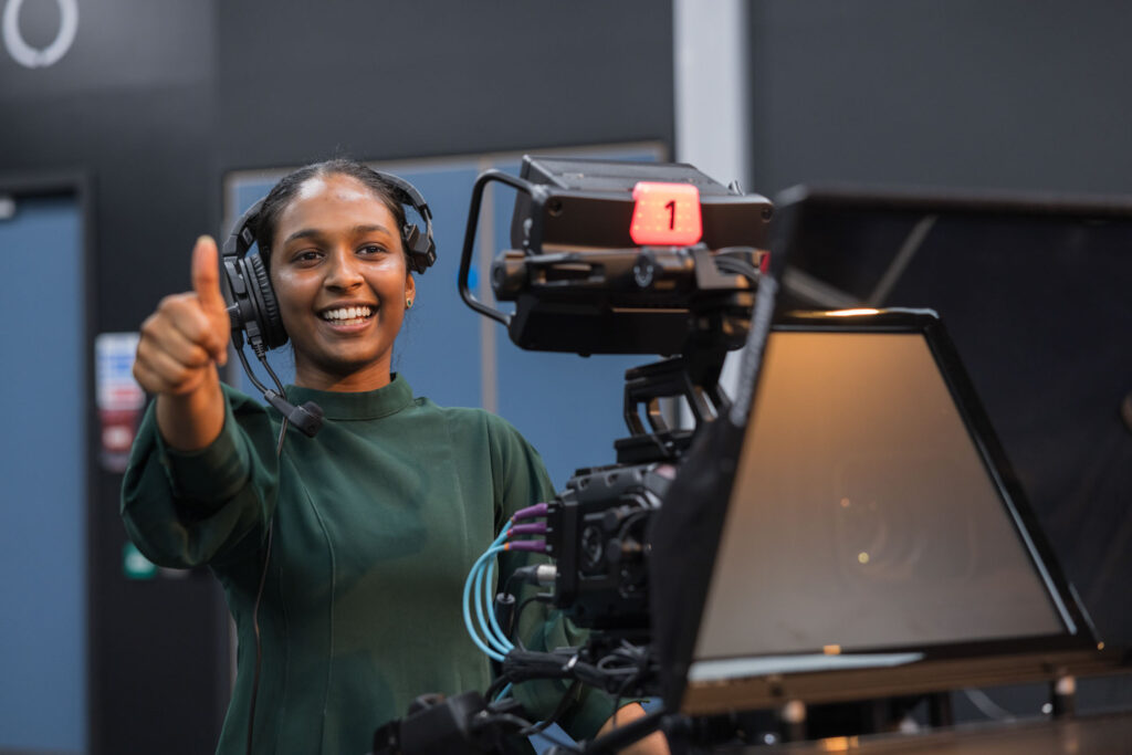 Student operating studio television camera giving a thumbs up signal.