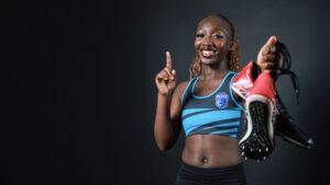 Female athlete holding running shoes, signalling number one with fore-finger.